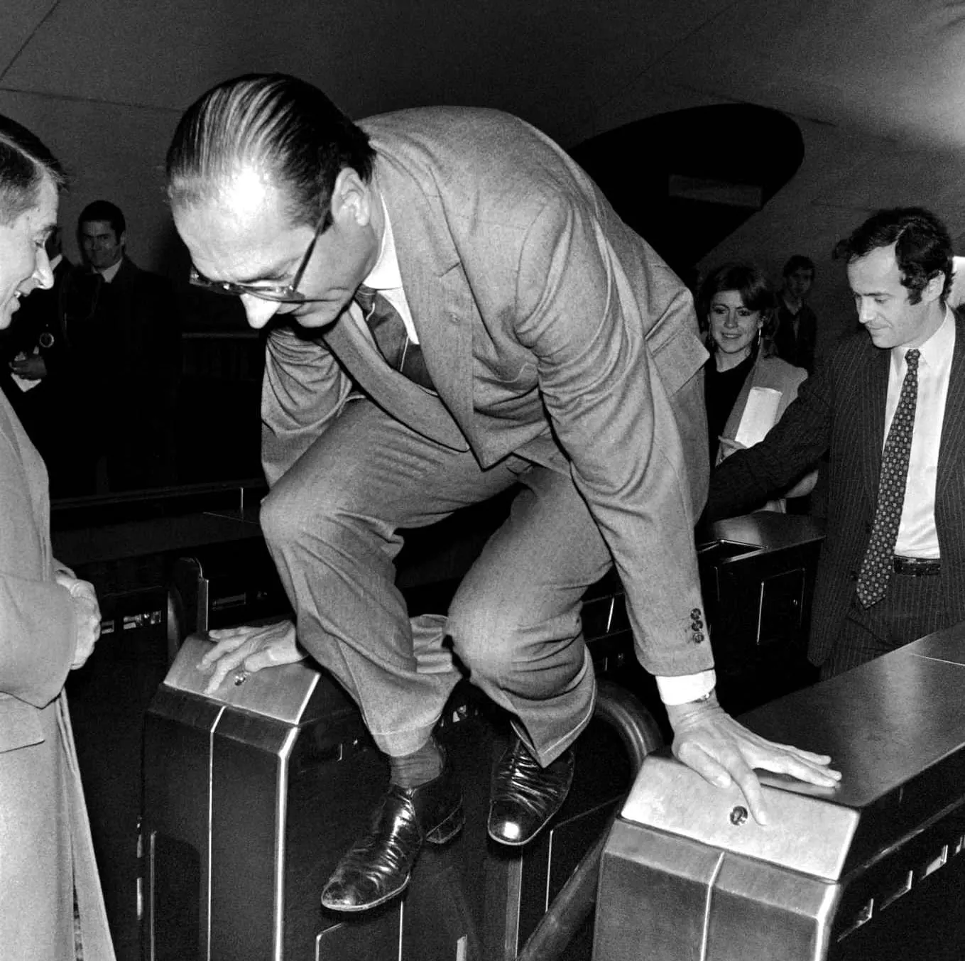 Black and white photograph of Jacques Chirac, mid-jump as he vaults over a metro gate; capturing a candid moment in a formal suit.